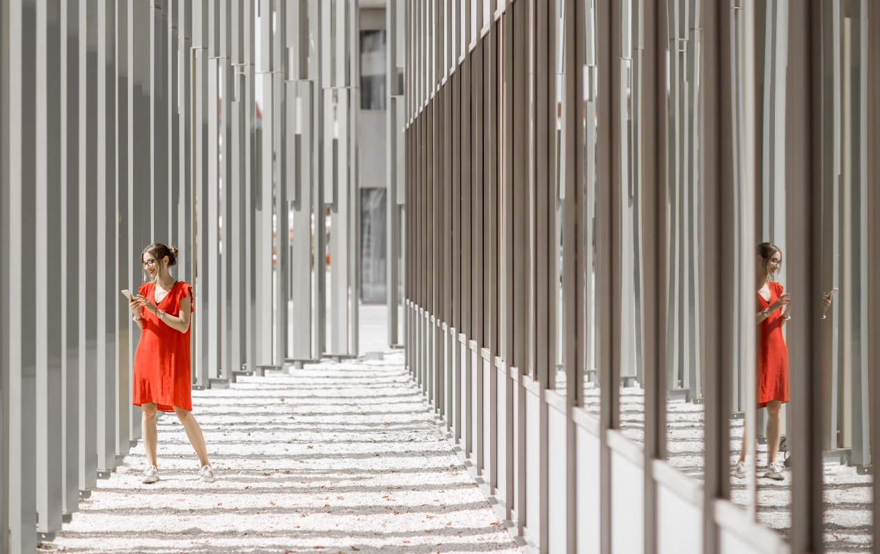 Woman in an orange-red dress holding a phone, standing in a passageway with architectural structures on both sides.