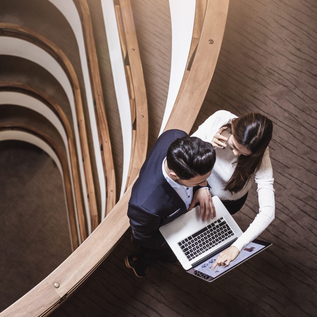 Bird's-eye view of a woman and man standing in a modern stairwell, looking at an open laptop screen together; a spiral staircase with a wooden railing is visible.